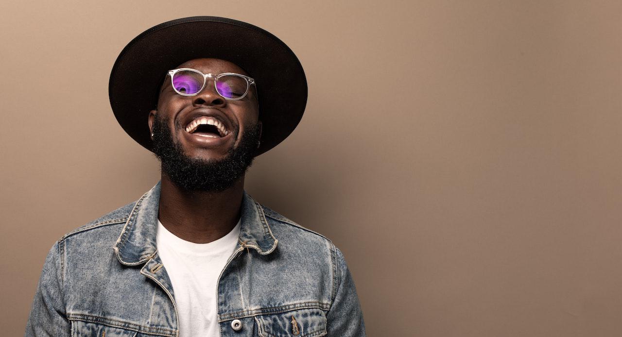 Black man in jean jacket, hat, and shades smiling at the camera